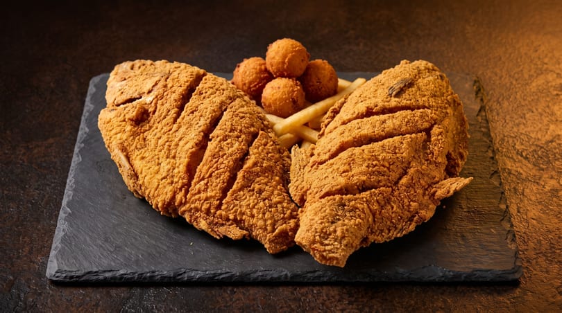 Two crispy fried fish fillets with french fries and hush puppies on a dark slate board.