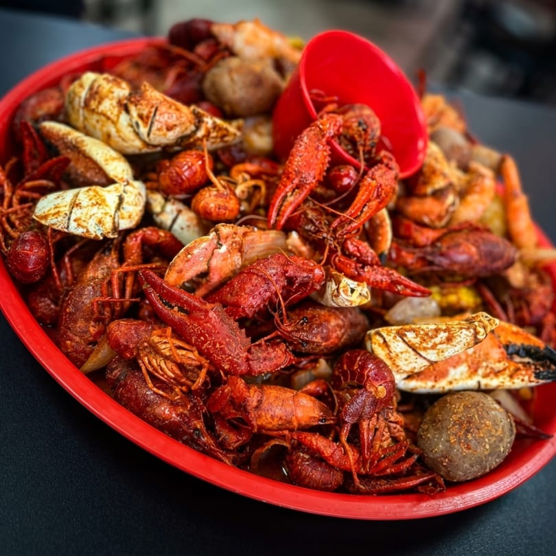 A red platter piled high with seasoned crawfish, crab legs, and potatoes, with a red cup.