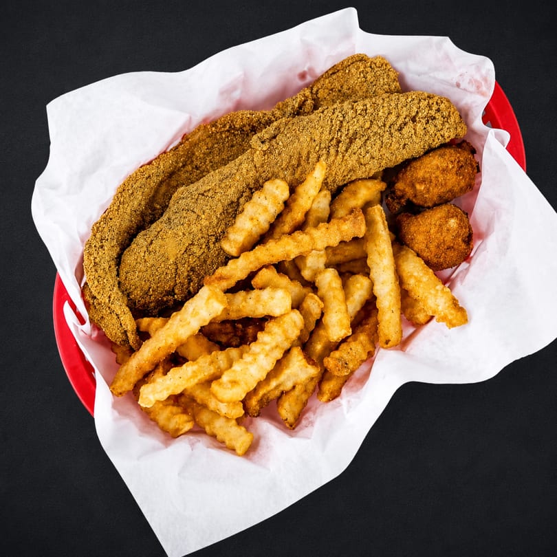 Fried fish fillets, crinkle-cut fries, and hush puppies in a red basket lined with white paper.