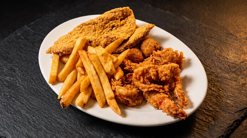 A white plate filled with golden-brown fried fish, french fries, hush puppies, and fried shrimp.