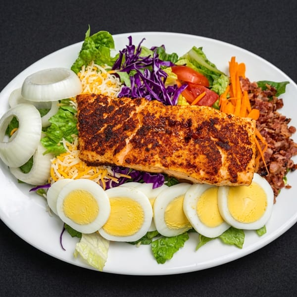 A grilled salmon salad with lettuce, cheese, onions, tomatoes, carrots, bacon bits, and hard-boiled eggs.