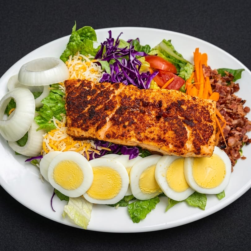 A grilled salmon salad with lettuce, cheese, onions, tomatoes, carrots, bacon bits, and hard-boiled eggs.